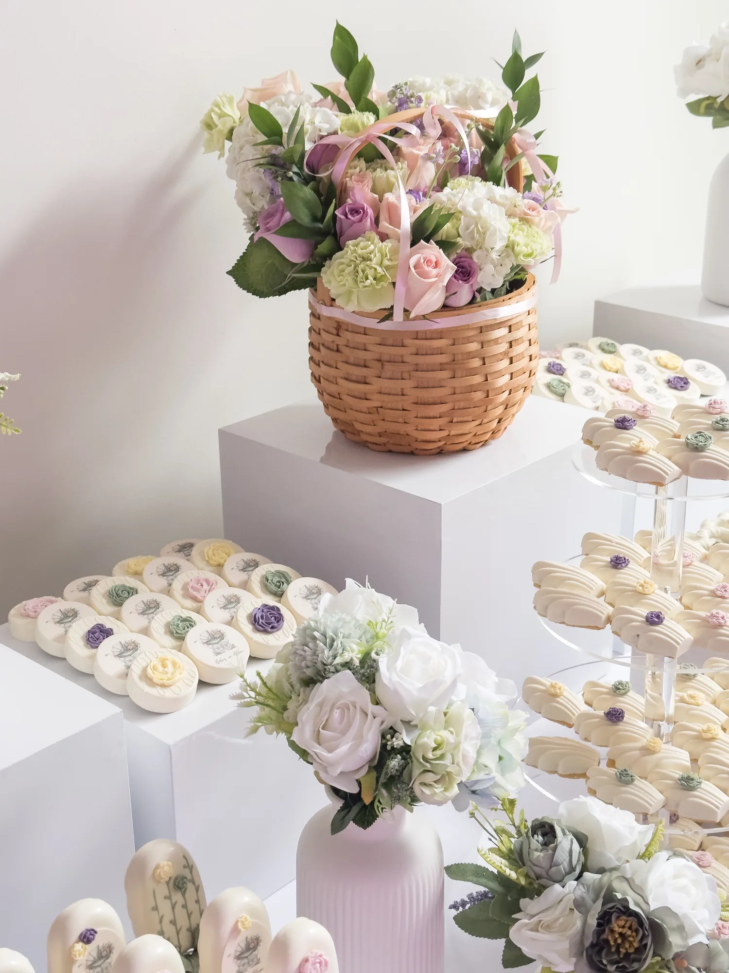 Printed chocolate covered Oreos with pastel sugar roses and tiered madeleines, beside a basket of fresh flowers.