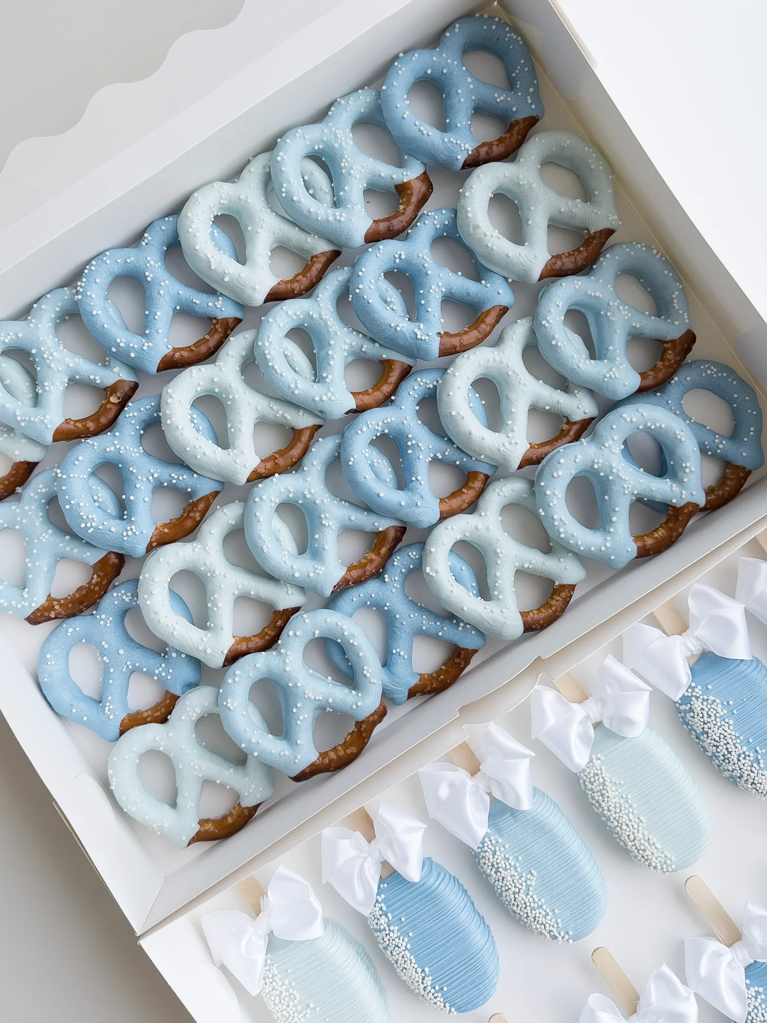 Rows of light blue and white chocolate dipped pretzels with white sugar pearls and small bows.