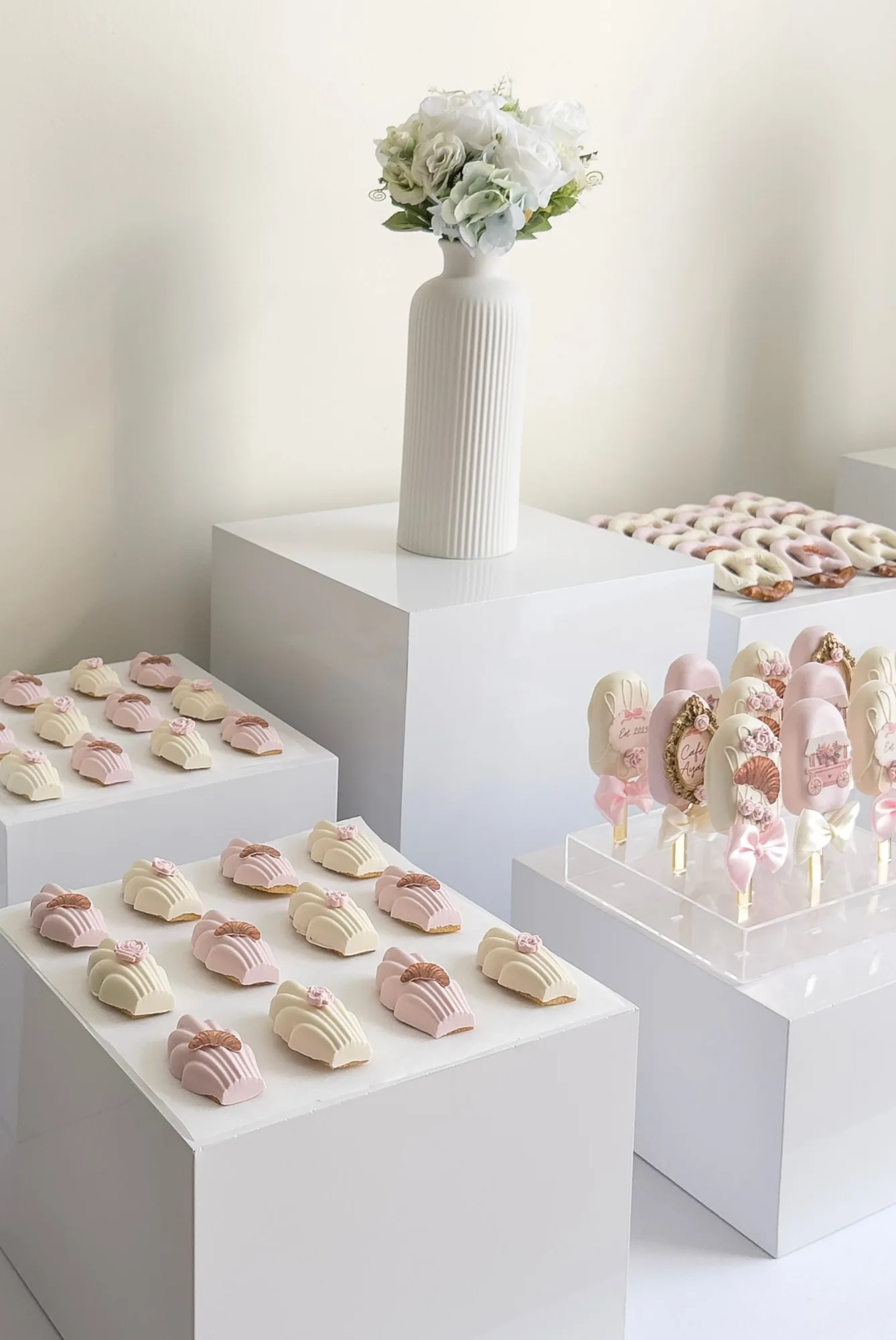 Pink and ivory chocolate covered madeleines on white display cubes, with a fluted vase of white flowers.