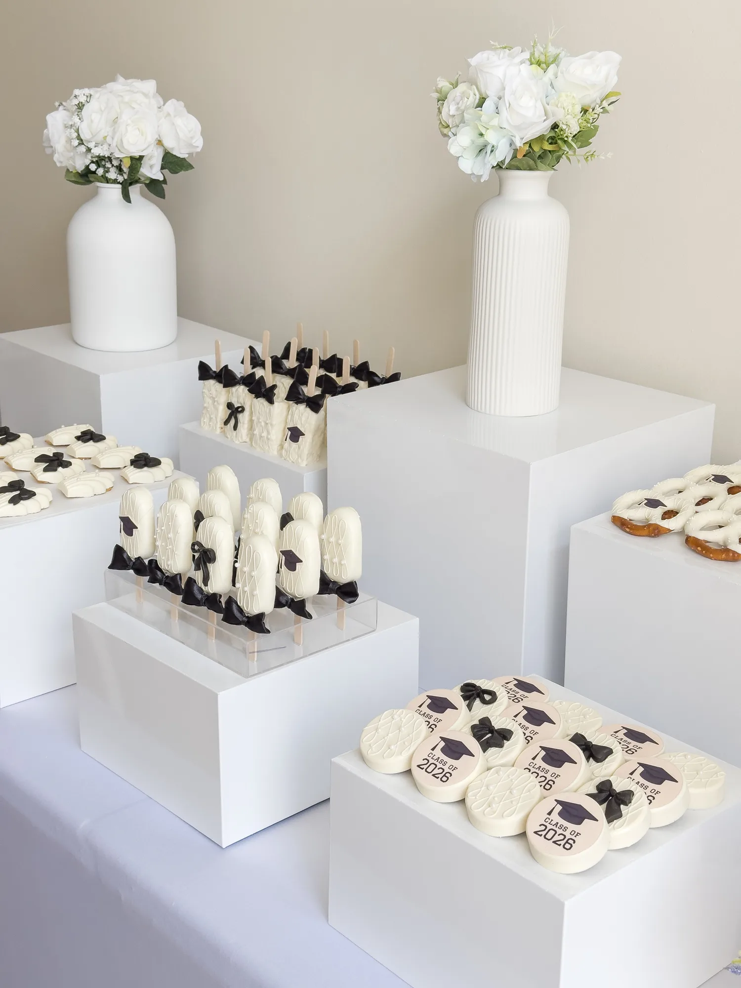White chocolate covered Oreos printed with Class of 2026 and black sugar bows, on white display cubes with white roses.