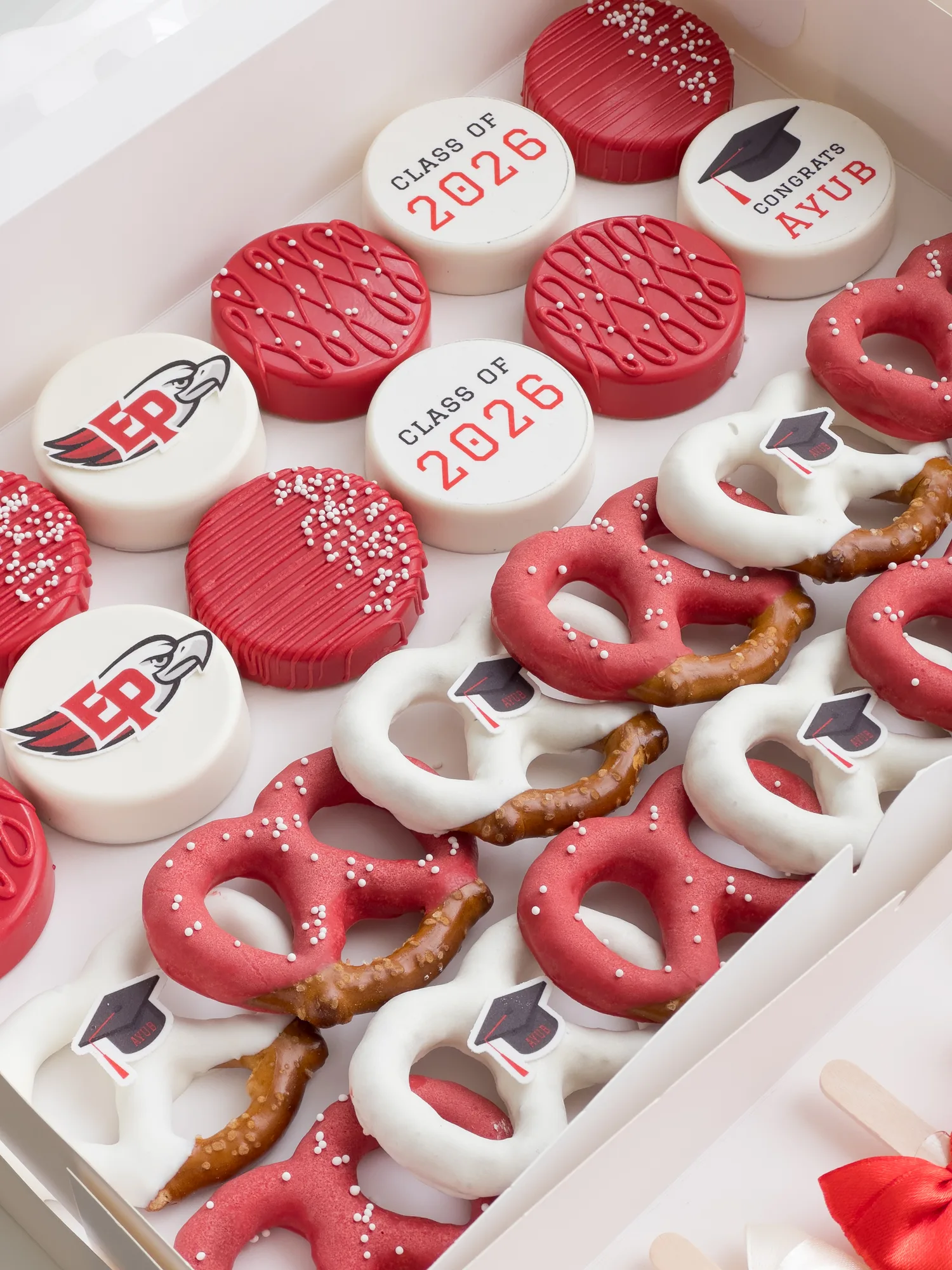 An open box of red and white Class of 2026 covered Oreos and dipped pretzels with black sugar graduation caps.