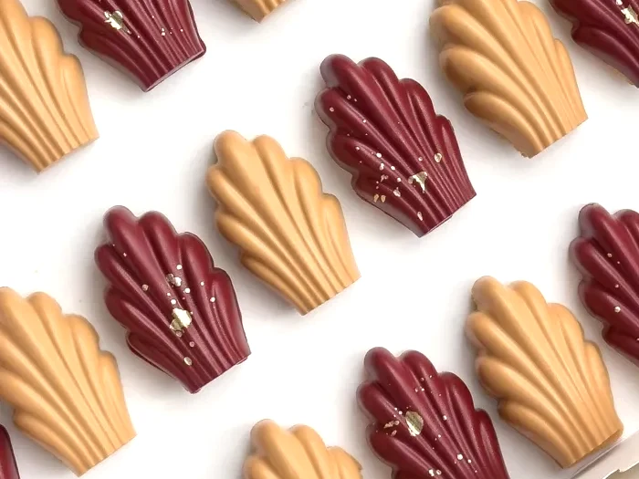 Caramel and burgundy madeleines flecked with gold, lined up in a white box.