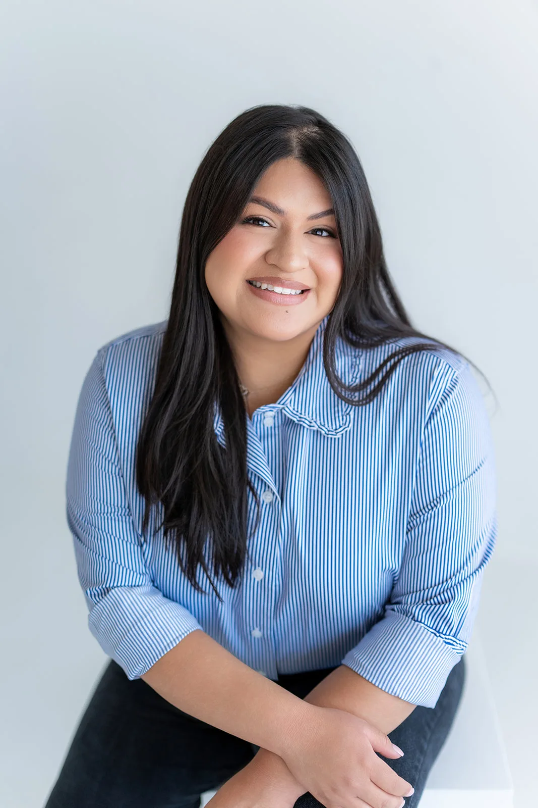 Maria, the owner of The Sweet Mission, seated in a blue striped shirt with her hands folded on her knee, smiling at the camera.