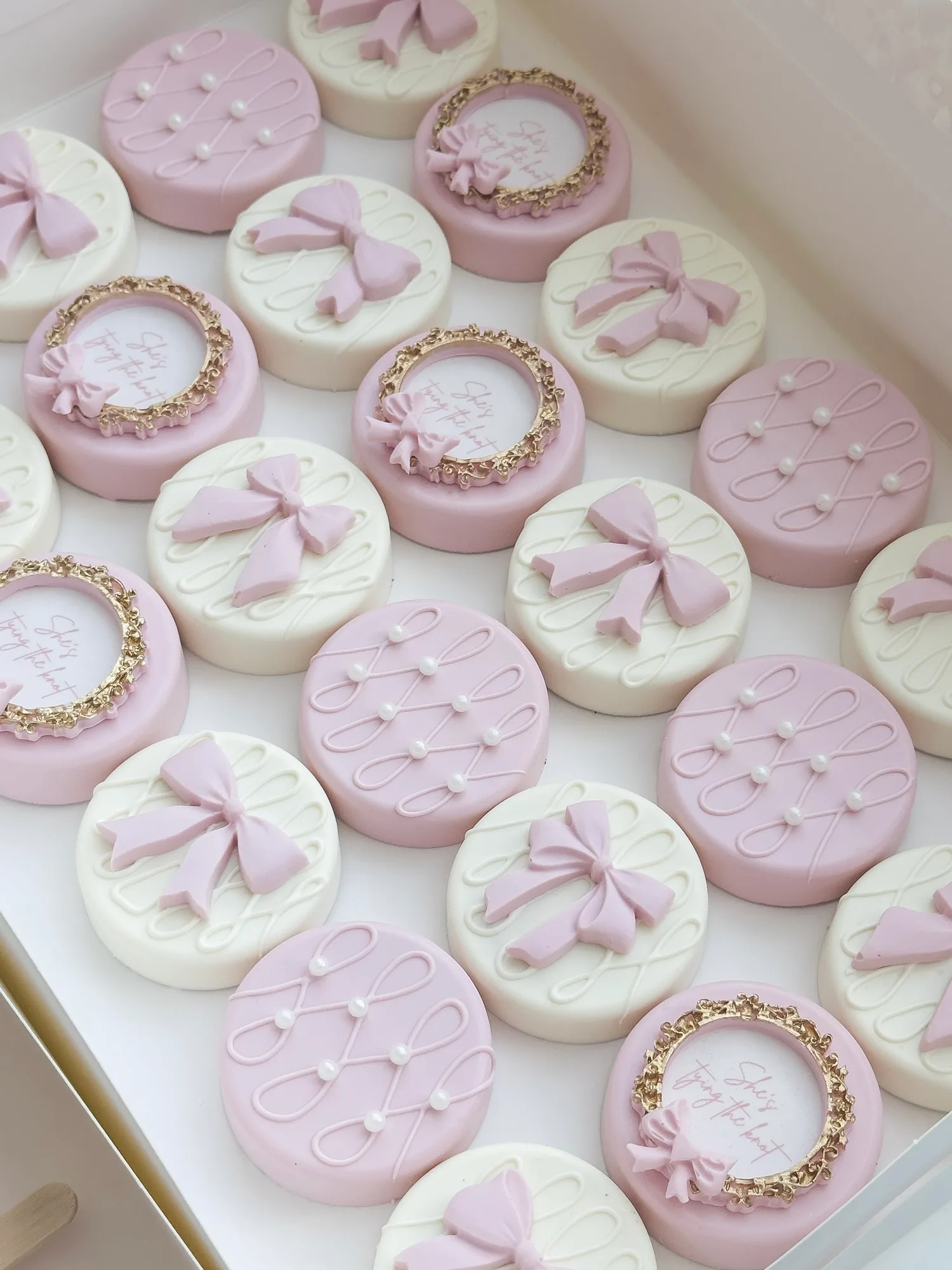 Pink and white covered Oreos topped with piped bows, sugar pearls, and gold framed plaques, packed in a white box.