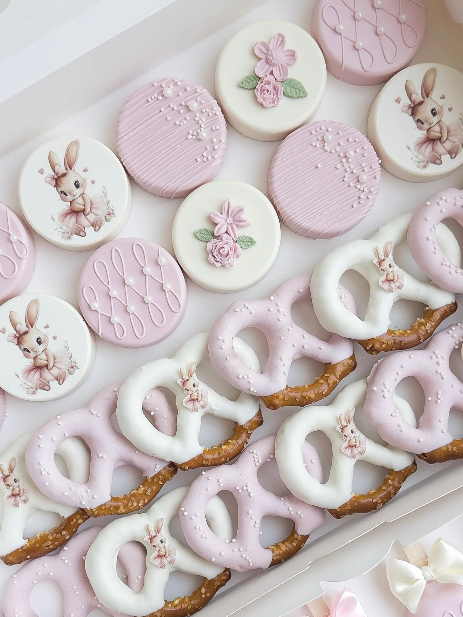 Pink and white baby shower treats: covered Oreos with a printed bunny and small pink flowers, beside pink and white dipped pretzels.