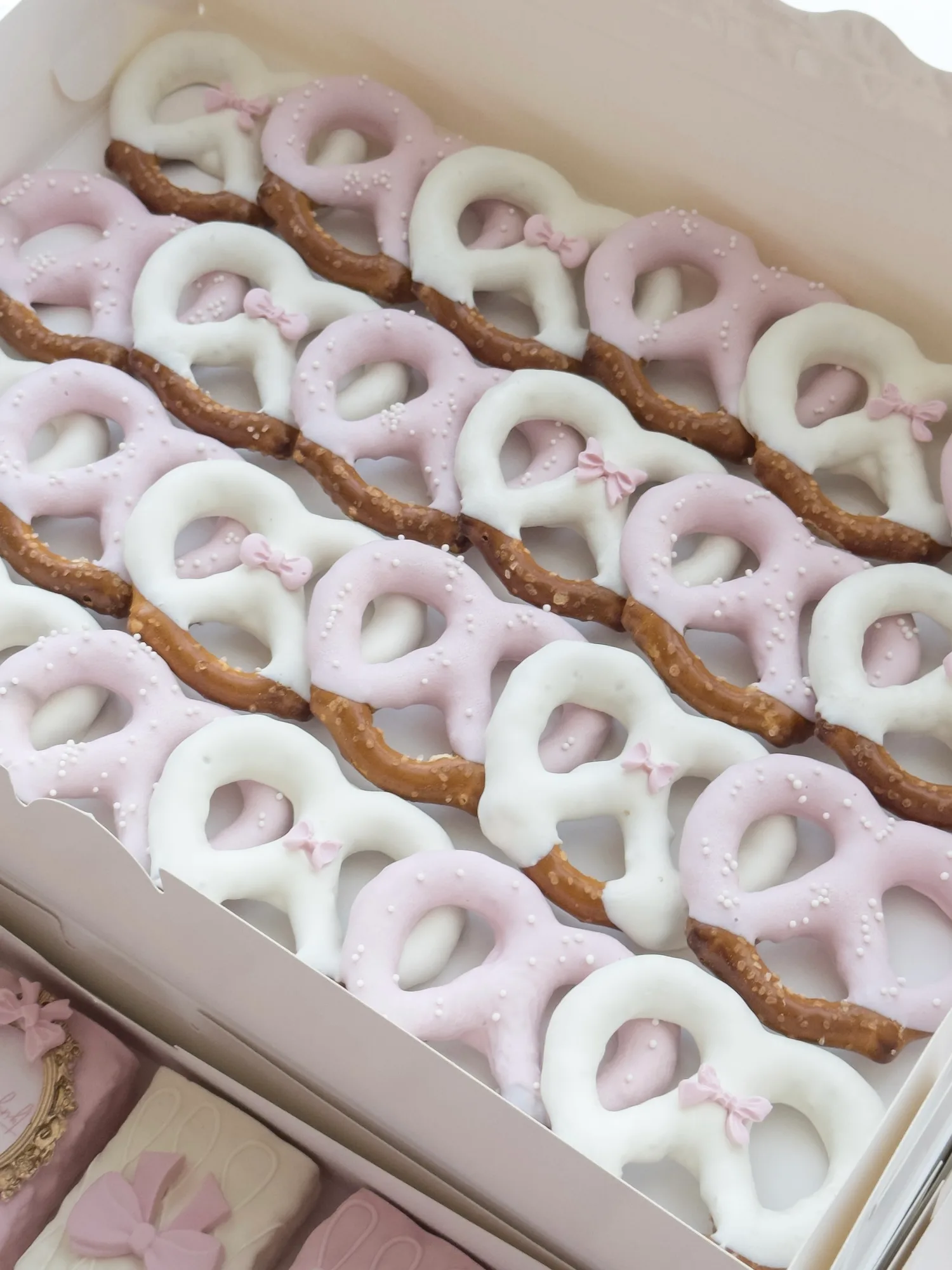 Rows of pink and white chocolate dipped pretzels with small piped bows, packed in a white box.