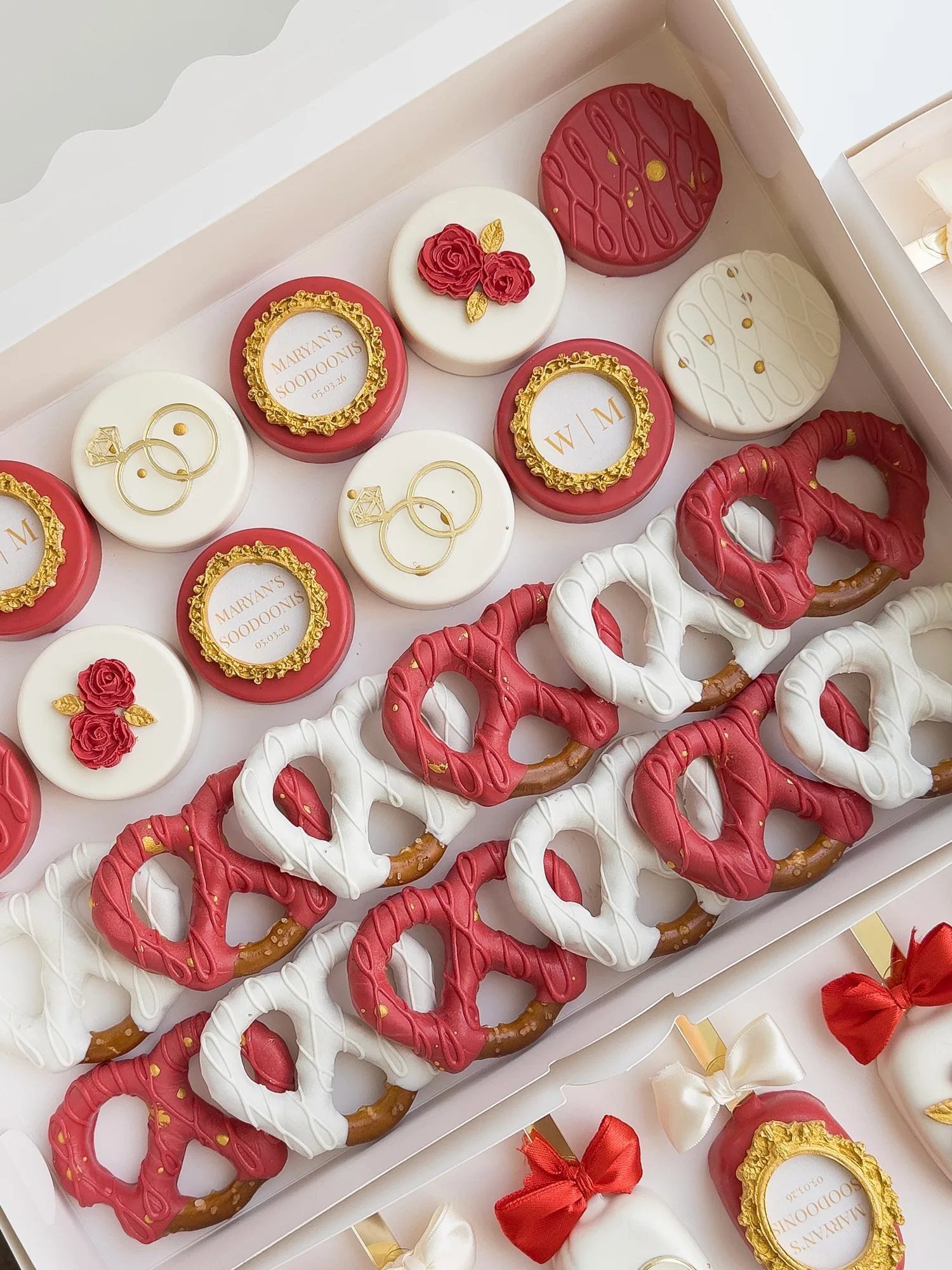 Red and ivory covered Oreos with gold rings, roses, and gold framed plaques, beside red and white dipped pretzels.