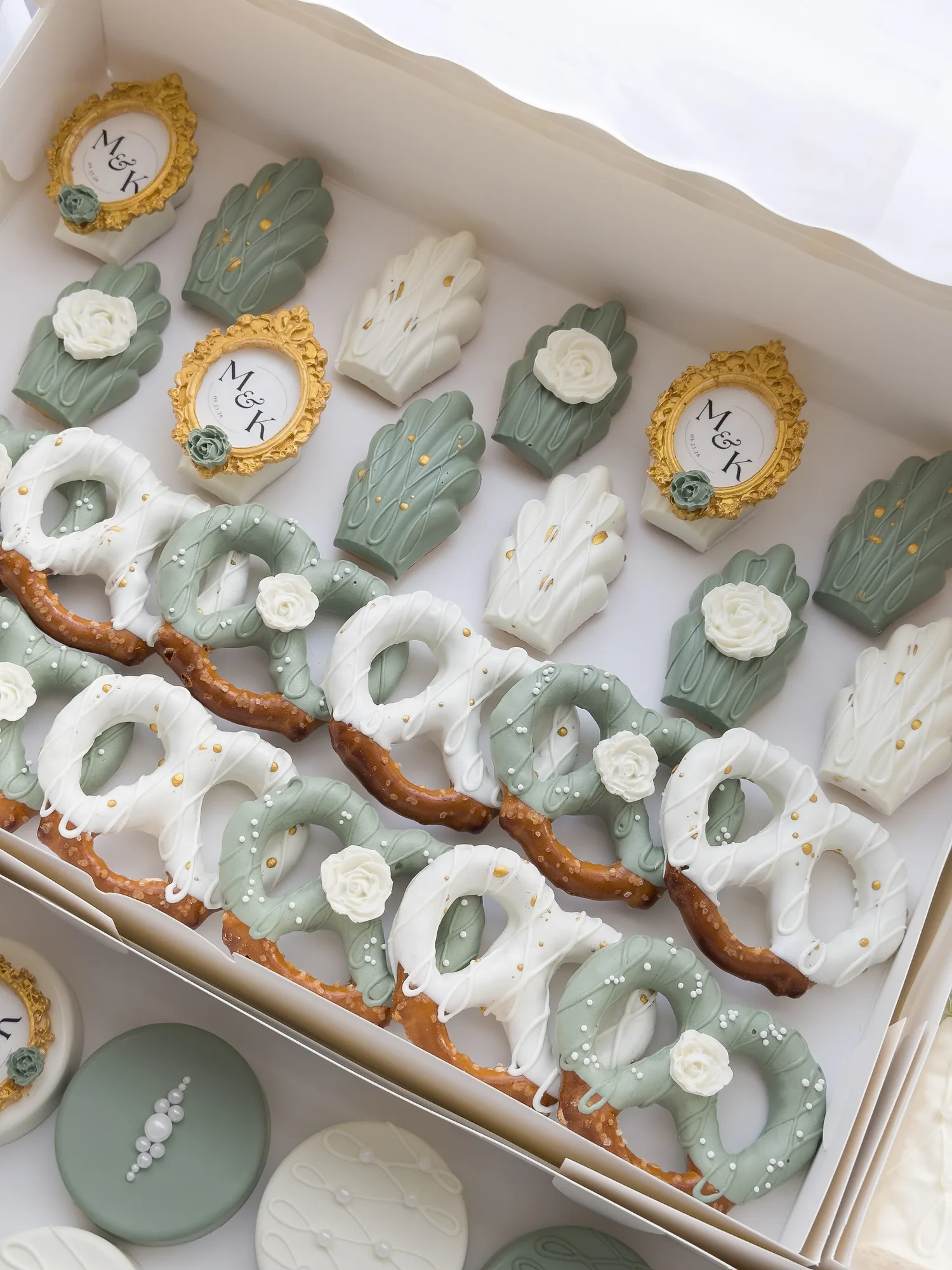 An open box of sage green and ivory treats with gold monogram frames, madeleines, and white dipped pretzels.