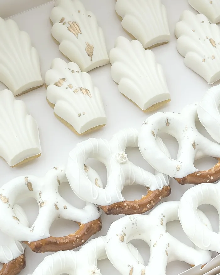 White dipped madeleines flecked with gold leaf, beside white pretzels finished with small sugar flowers.